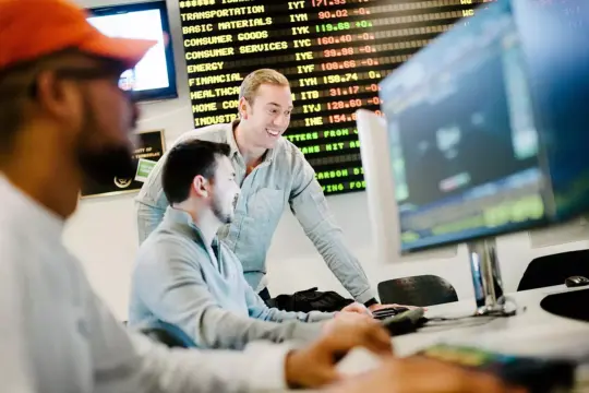 Three people in an office setting focus on computer screens displaying stock market data. A man in a light shirt stands, interacting with two seated colleagues. A digital board in the background shows various financial figures and categories.