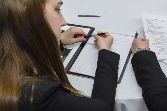 A person with long brown hair writes in a notebook at a table. Another person sits beside them, holding a sheet of paper. Both wear black suits, and a pen and a notebook are on the table.