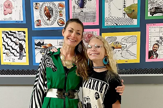 Two people stand smiling with arms around each other in front of a colorful wall display featuring various artwork. The person on the right, wearing glasses and a Nirvana shirt, celebrates their involvement in the Teacher Intern Program.