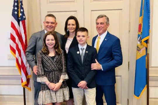 A group of five people, including two men in suits, a woman, and two children, one boy and one girl, standing together in a room with flags on either side. The setting appears formal, and everyone is smiling.