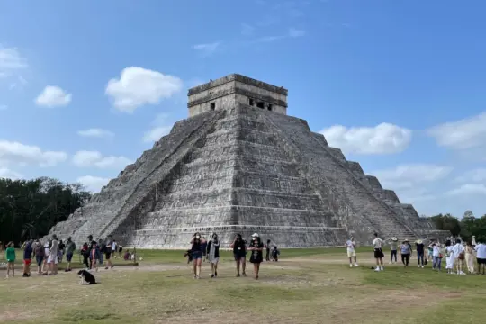 A large stone pyramid with stepped sides rises against a blue sky with clouds. Numerous people walk and gather around the base on grassy ground. Trees line the horizon.