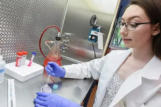 A scientist in a lab coat and gloves is handling a petri dish with a pipette. Test tubes and a flask are on the counter beside her. She is working in a laboratory environment.