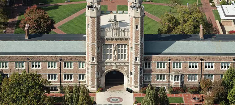 Aerial view of a large, historic stone building with towers and archway, surrounded by trees and greenery on a college campus. Paths are visible on a lawn behind the building.