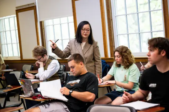 A teacher stands and points while students sit at desks in a classroom. Some students take notes, while others look at the teacher. Light streams in through large windows, and a laptop is open on one student's desk.