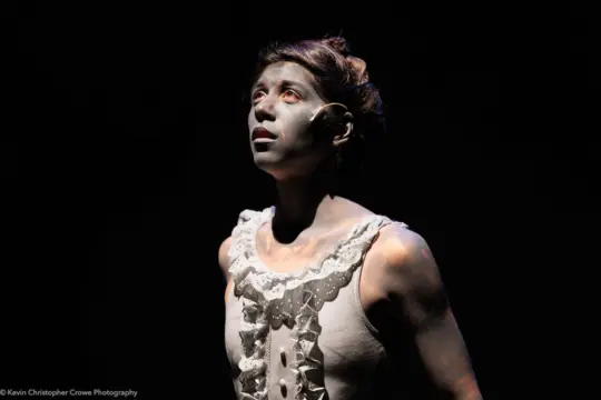 A person with their face and upper body painted in gray makeup stands on stage in dramatic lighting, wearing a sleeveless garment with lace and button details. They look upwards with an expression of awe or contemplation.