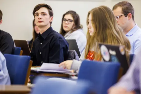 Students sitting in a classroom, attentively listening and taking notes. Some have laptops open, while one person is focused on a stack of papers. The atmosphere is studious and engaged.