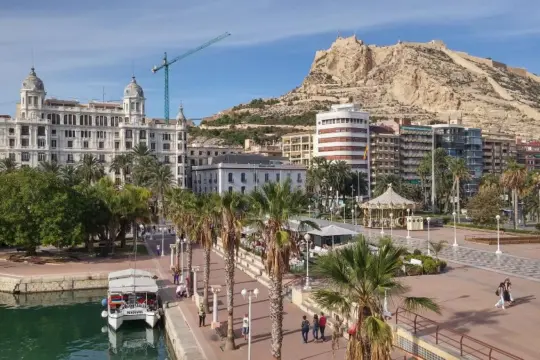 Scenic view of Alicante, Spain, featuring the Santa Bárbara Castle atop a hill. In the foreground, there's a marina with a docked boat, palm trees, and a paved promenade with people walking. Buildings line the background under a clear blue sky.