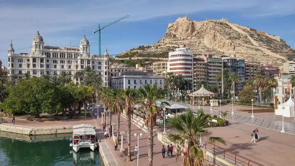 Scenic view of Alicante, Spain, featuring the Santa Bárbara Castle atop a hill. In the foreground, there's a marina with a docked boat, palm trees, and a paved promenade with people walking. Buildings line the background under a clear blue sky.