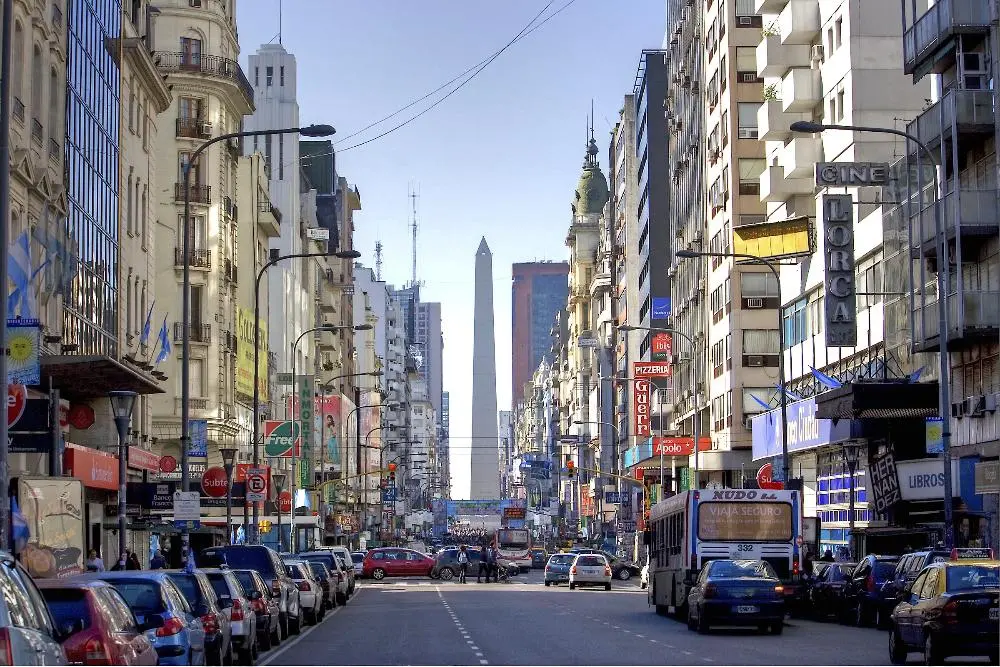 A wide street in a busy city lined with buildings and shops. Cars and buses fill the street. In the distance, a tall obelisk stands centrally. Bright blue sky overhead and various signs and advertisements are visible along the buildings.