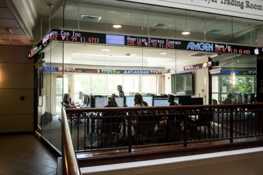 A group of people work on computers in a glass-enclosed room displaying various stock tickers. The room is surrounded by scrolling digital stock prices and company names such as Amgen, American Express, and Atlassian.