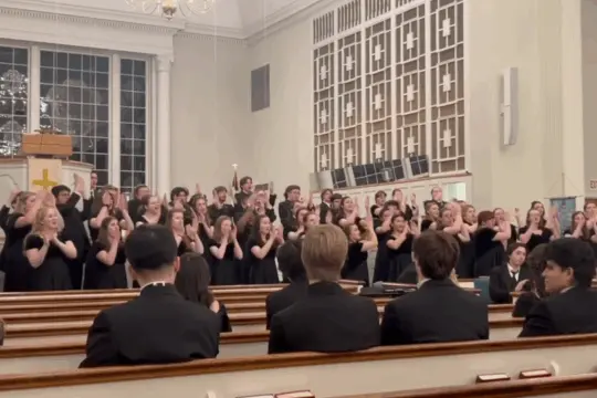 A choir of students dressed in black sings and gestures enthusiastically at the front of a church during a vibrant music performance, while audience members seated in pews watch. The church features tall windows and a large decorative organ in the background.