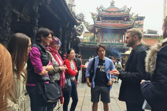 A group of people stands in conversation near a traditional temple with ornate architecture and incense burning. The scene, reminiscent of an 亚洲研究 discussion, appears lively and engaged against the backdrop of the cultural landmark.