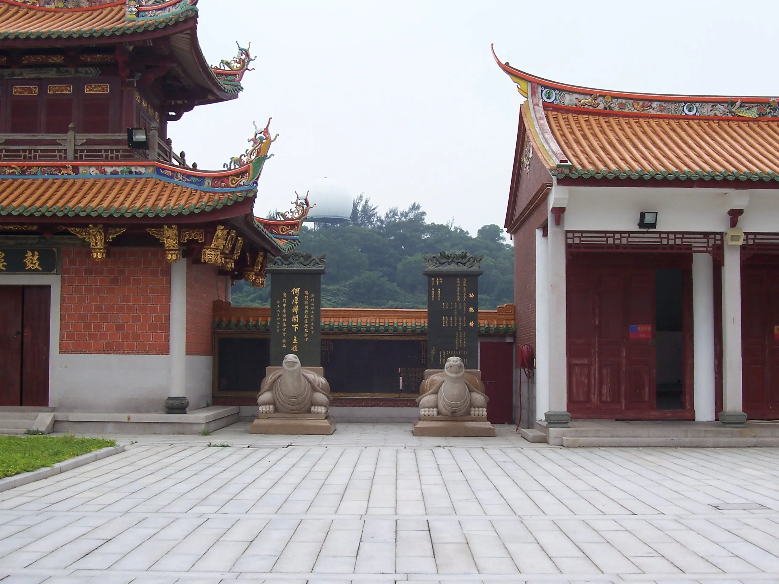 A traditional Chinese temple with ornate red and gold roofs, featuring detailed carvings reminiscent of applied linguistics. Two stone statues sit to the left and right of the entrance, resembling ancient guardians. The background shows trees under a cloudy sky.