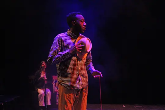 A theater performance of a man in a checkered shirt holds a hat and a cane on a dimly lit stage. He stands in the foreground with a thoughtful expression. In the background, a person sits on a chair, partially obscured by shadows.