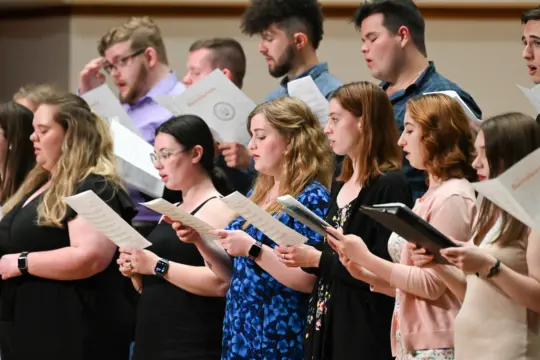 A diverse choir group rehearses on stage, holding music sheets and performing church music. The singers, both men and women, stand in rows, focused and engaged in their harmonious performance. The setting is a well-lit indoor space.