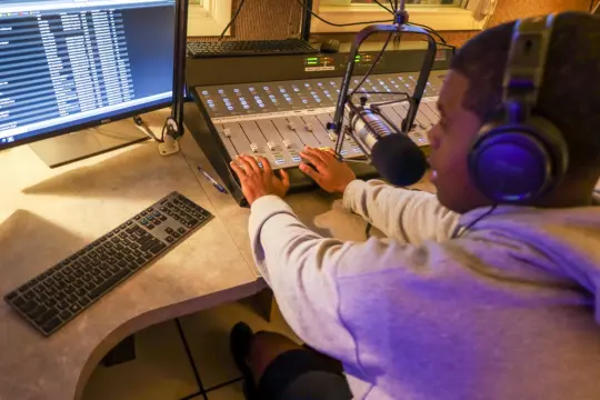 A person wearing headphones operates a soundboard in the warmly lit recording studio, engaging in the art of communication. They speak into a microphone, while a computer monitor displays audio software nearby, emphasizing the seamless fusion of technology and creativity in audio production work.