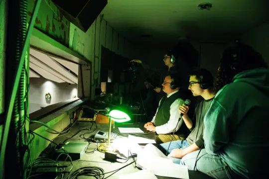 In a dim control room, where the art of communication studies comes to life, people focus on broadcasting an event. One operates the camera while others speak into microphones. Their faces are lit by the green glow of a desk lamp, illuminating sheets of paper and equipment cluttering the desk.