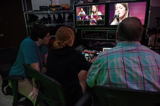 Three people in a control room sit at a desk with multiple monitors displaying a woman singing. Focused on managing audio and video equipment, they utilize communication studies principles to enhance the live feed. Shelves with additional gear are in the background.