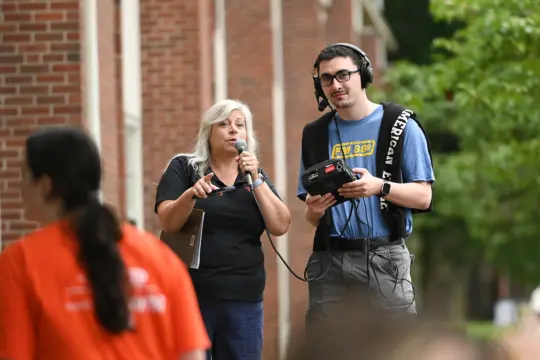 A woman with long gray hair speaks into a microphone outdoors, embodying the essence of Communication Studies. Beside her, a man wearing glasses and headphones holds recording equipment near a brick building, framed by trees. A blurred figure in the foreground adds depth to this collaborative scene.