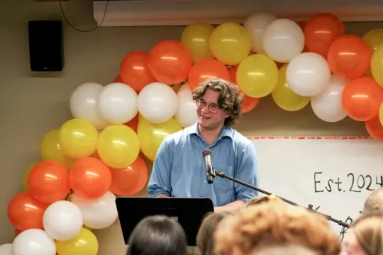 A person in a blue shirt, immersed in the art of creative writing, speaks at a podium with a microphone, smiling. Behind them, a vibrant balloon arch of orange, yellow, and white bursts with color. In the background, 