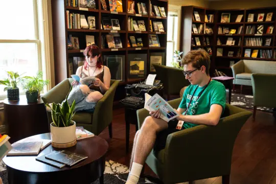 Two people are sitting in a cozy reading room with bookshelves. One is on the left in an armchair, engrossed in creative writing while holding a plush toy. The other, wearing a green shirt, is reading a book on the right. A typewriter and plants are scattered around them.