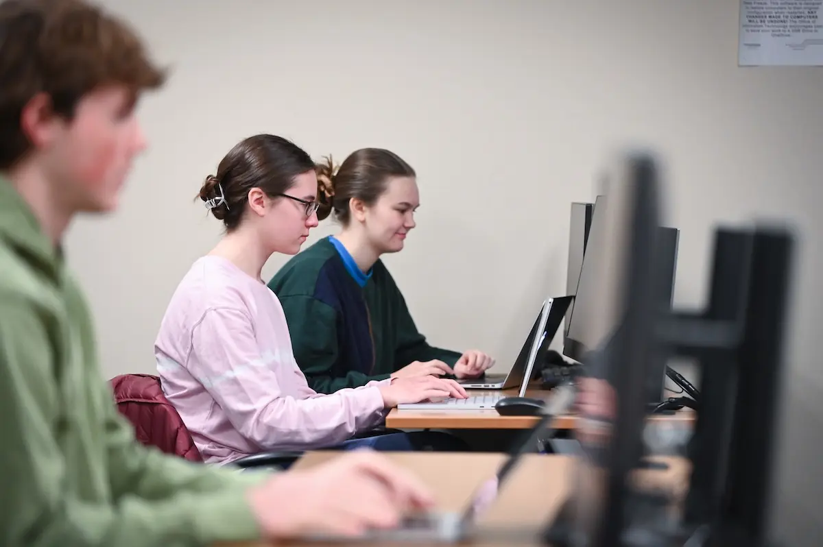 Three people sit side by side at a row of desks, focused on their computers. They work intently in a classroom setting. The background is plain, and lighting is bright.