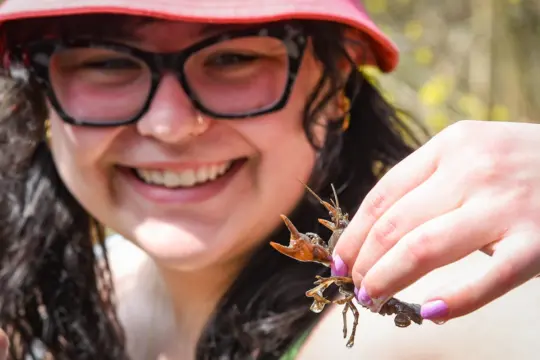 A person wearing a red hat and glasses, smiling and holding a twig with a small insect. The focus is on the insect, with the person's face slightly blurred in the background.
