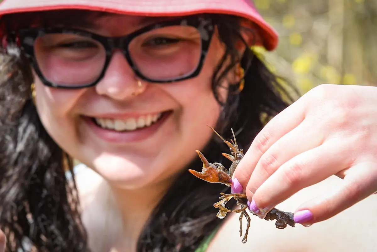 A person wearing a red hat and glasses, smiling and holding a twig with a small insect. The focus is on the insect, with the person's face slightly blurred in the background.