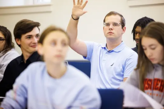 A man in a light blue polo shirt raises his hand in a classroom. Several other students are seated around him, some are looking at him, while others are focused on their work. The setting is an economics lecture or discussion session.