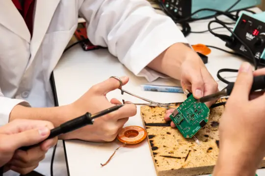 Three people collaborate on circuit board assembly at a table, integrating skills from the Engineering 3+2 Program. One holds the board with pliers as another uses a soldering iron, while a third person assists. Tools and equipment are scattered around them, illustrating hands-on learning.