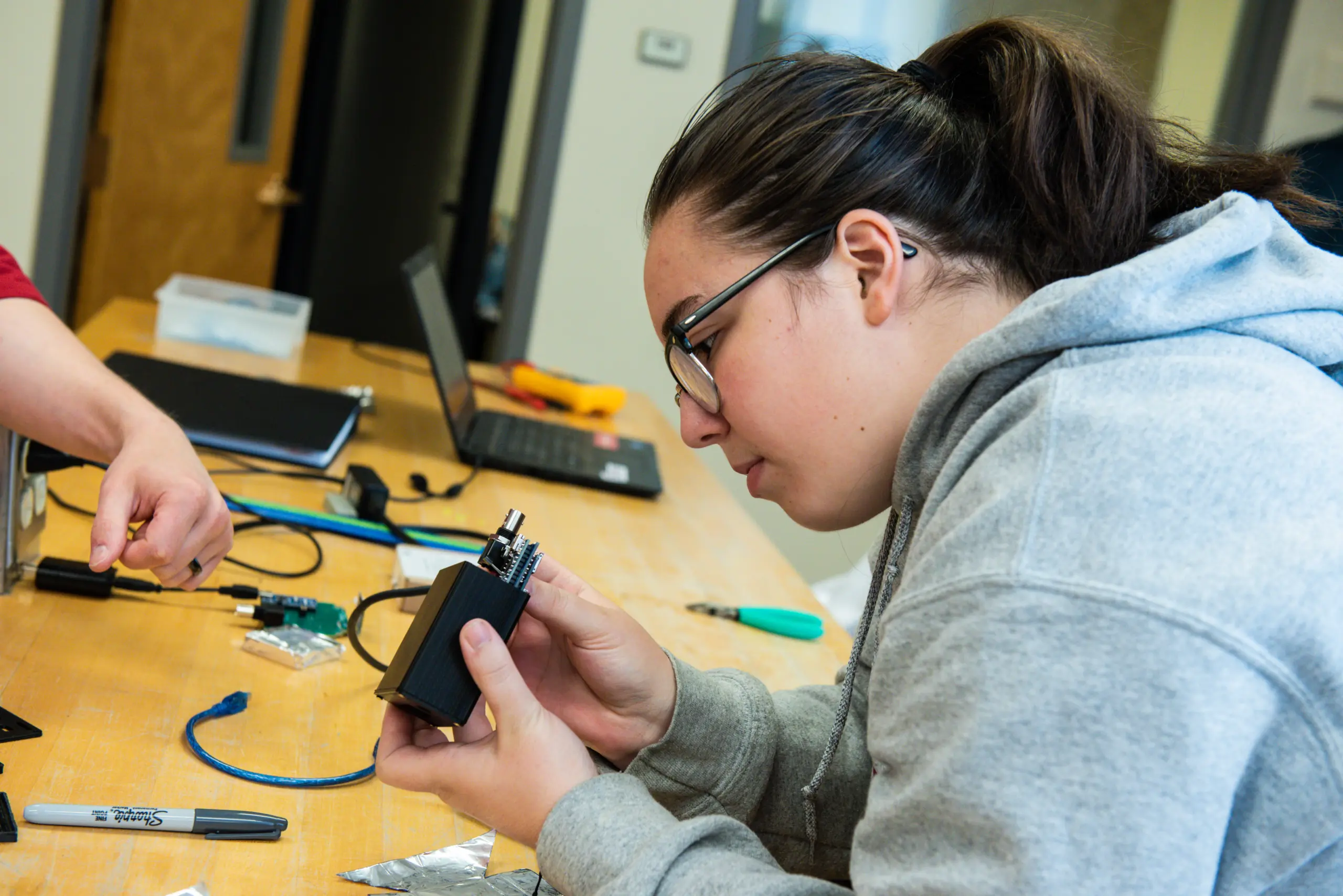 A person in a gray hoodie, possibly part of an Engineering 3+2 Program, examines an electronic device at a table with various tools and wires. Another person’s hand points toward the device. In the background, a laptop rests on the wooden table.