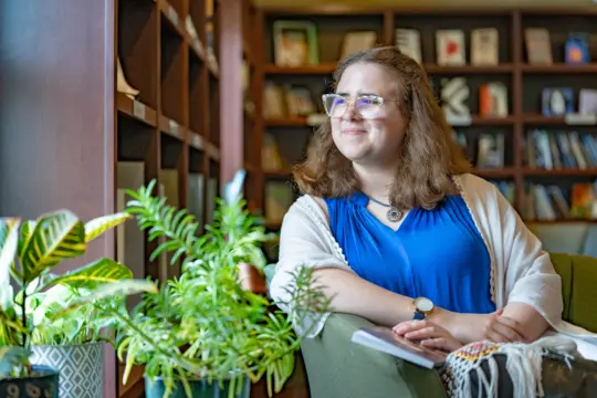 A person with glasses and long hair sits in a cozy library, perhaps pondering English literature as they gaze out the window. They wear a blue top and white shawl, with a watch on one wrist. The green potted plants and bookshelves enhance the tranquil atmosphere.