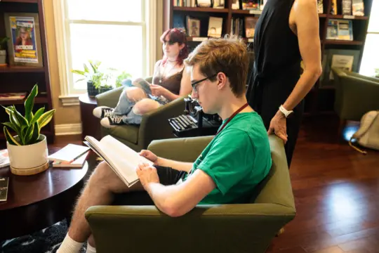 Two people are sitting in a library. A person in a green shirt reads an English literature book in the foreground, while another person with red hair reads in the background. A third person stands nearby. Bookshelves and plants decorate the room.