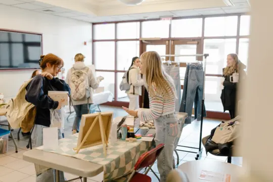 People are interacting at an indoor craft fair. A woman behind a table with crafts speaks with a visitor. Others browse items on display. A rack of clothing is visible in the background, and the atmosphere is casual and bright.