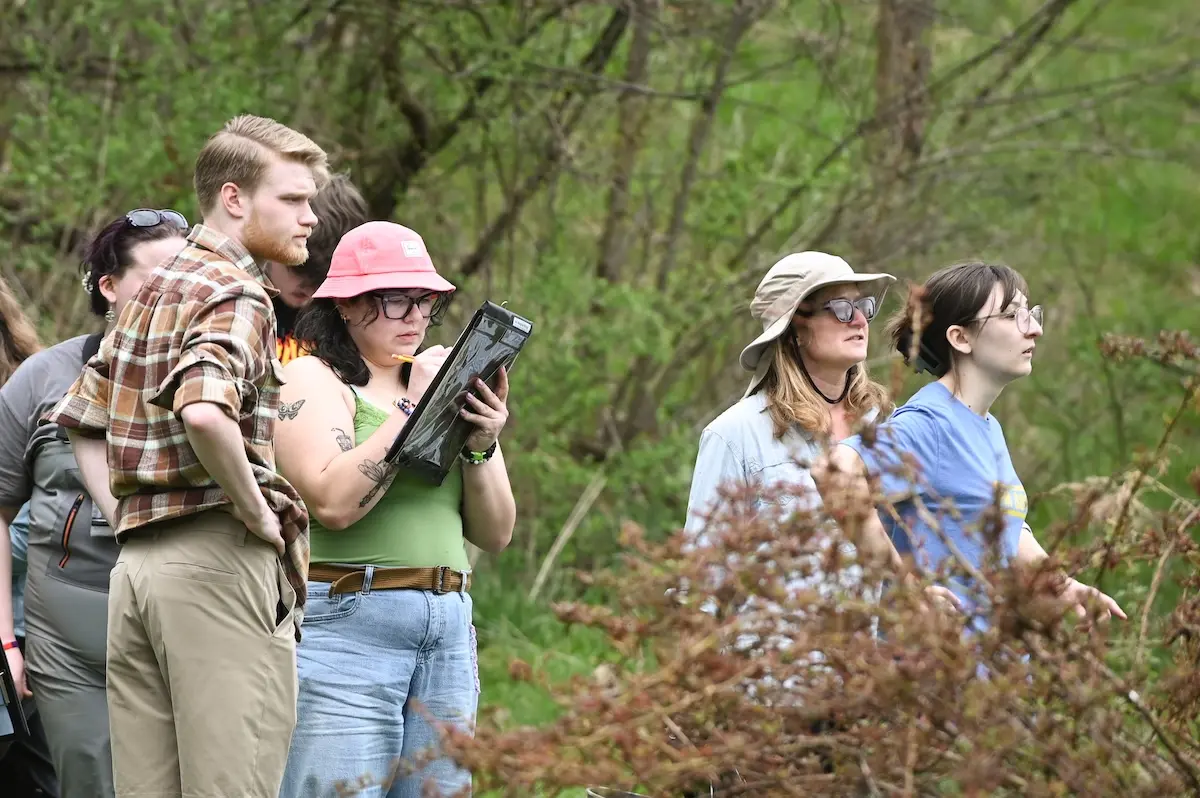 A group of people stand together outdoors, observing something off-camera. One person takes notes on a clipboard. They are surrounded by green foliage and overcast skies, dressed casually with hats for sun protection.