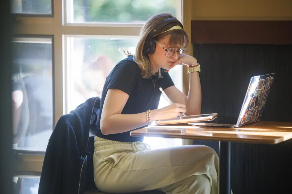 A person wearing glasses and headphones sits at a table in front of a laptop, jotting down notes on English Literature with a stylus on their tablet. They're dressed in a black shirt and light-colored pants, as sunlight streams in from the window behind them.