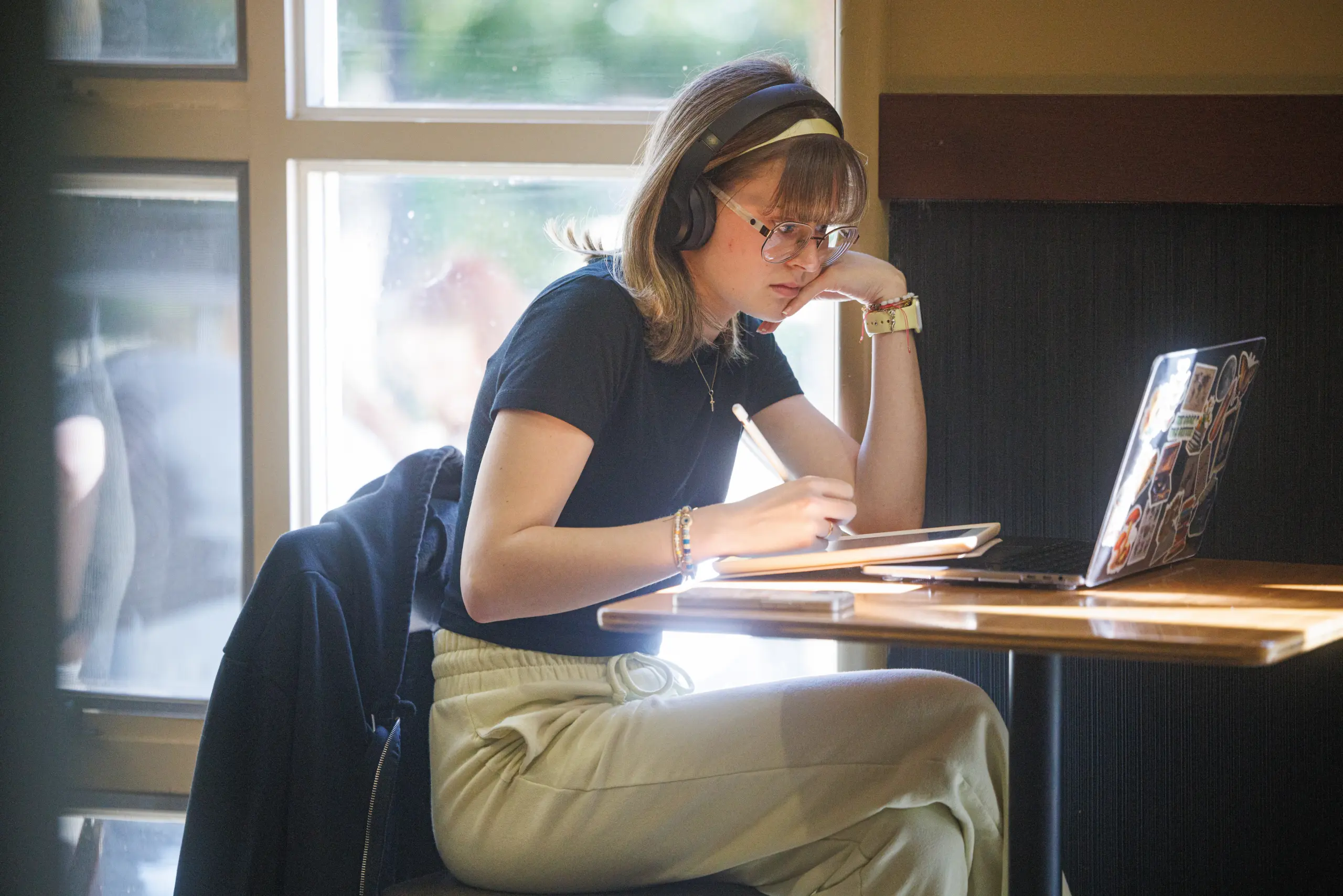 A person wearing glasses and headphones sits at a table in front of a laptop, jotting down notes on English Literature with a stylus on their tablet. They're dressed in a black shirt and light-colored pants, as sunlight streams in from the window behind them.