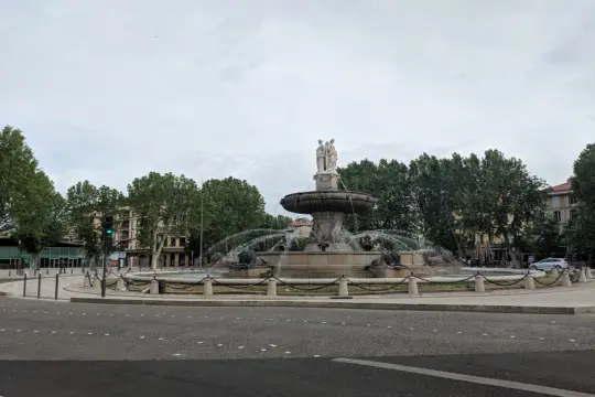 The image showcases a grand stone fountain with statues atop, reminiscent of classic French architectural elegance. Water arcs gracefully, surrounded by trees. This urban setting features buildings in the background under a cloudy sky, inviting thoughts akin to scenes from French studies.