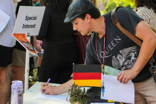 A person in a Queen T-shirt signs a paper at a table with a German flag. A 