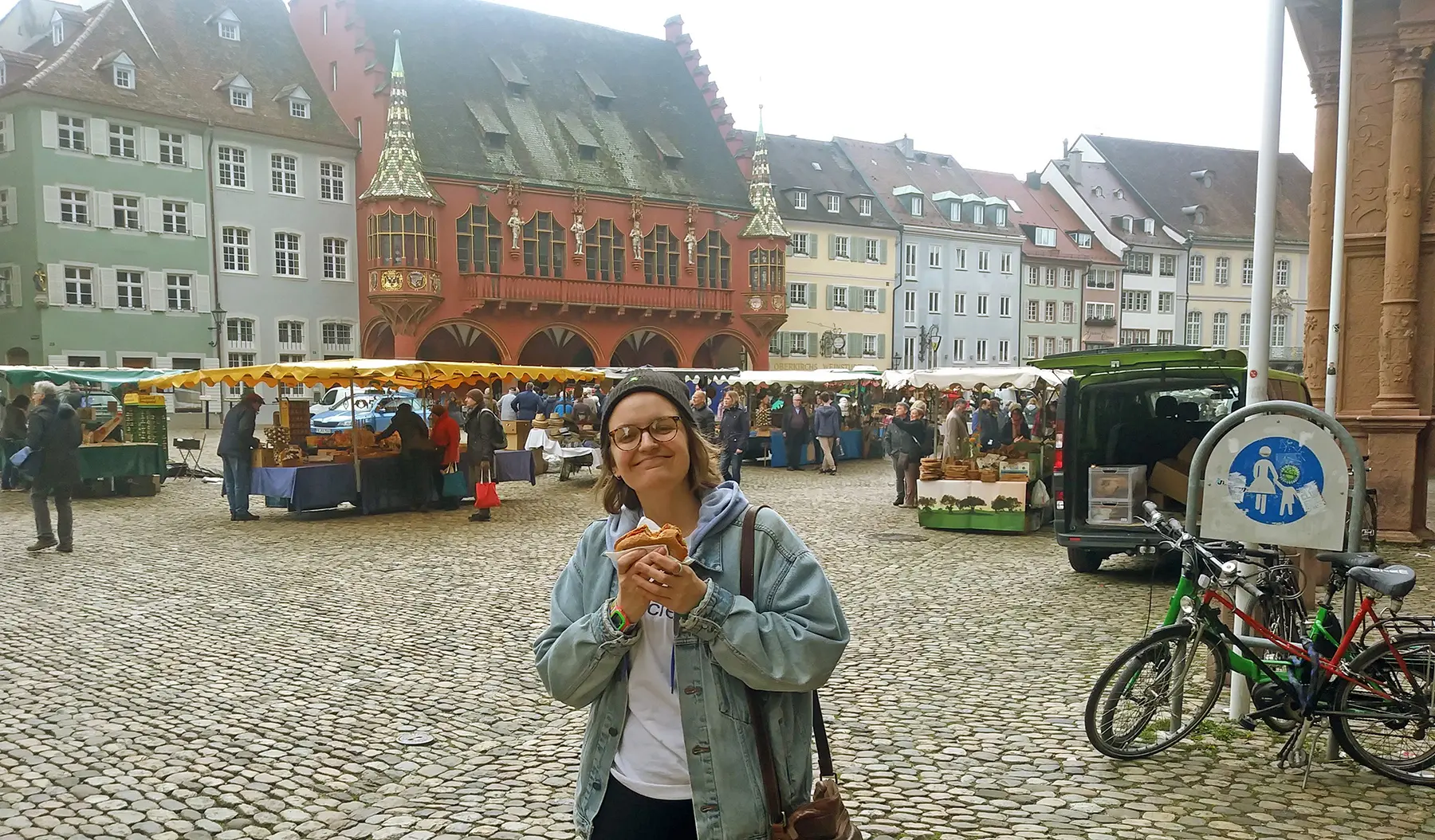 A person in a denim jacket and beanie stands smiling while holding food in a cobblestone square. Behind them, there's a market with stalls and people browsing. Colorful historic buildings and a red building with ornate details are visible.