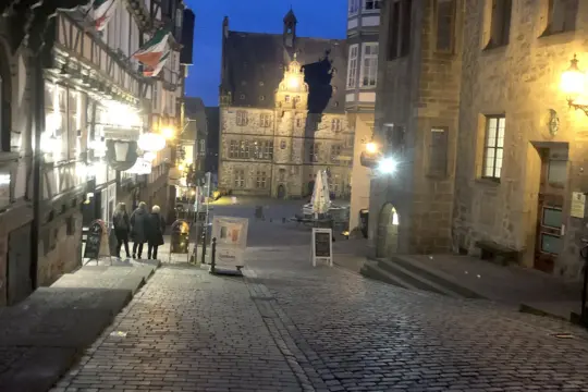 A cobblestone street in a historic town at dusk, with people walking downhill. The street is lined with old buildings, some with wooden beams. Soft lights illuminate the scene, and a large, ornate building stands at the end of the street.