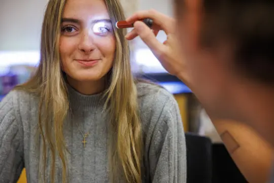 A person with long hair and a gray sweater sits while another person shines a small light into their eye, possibly during an eye examination. The person is smiling slightly, and a cross necklace is visible.