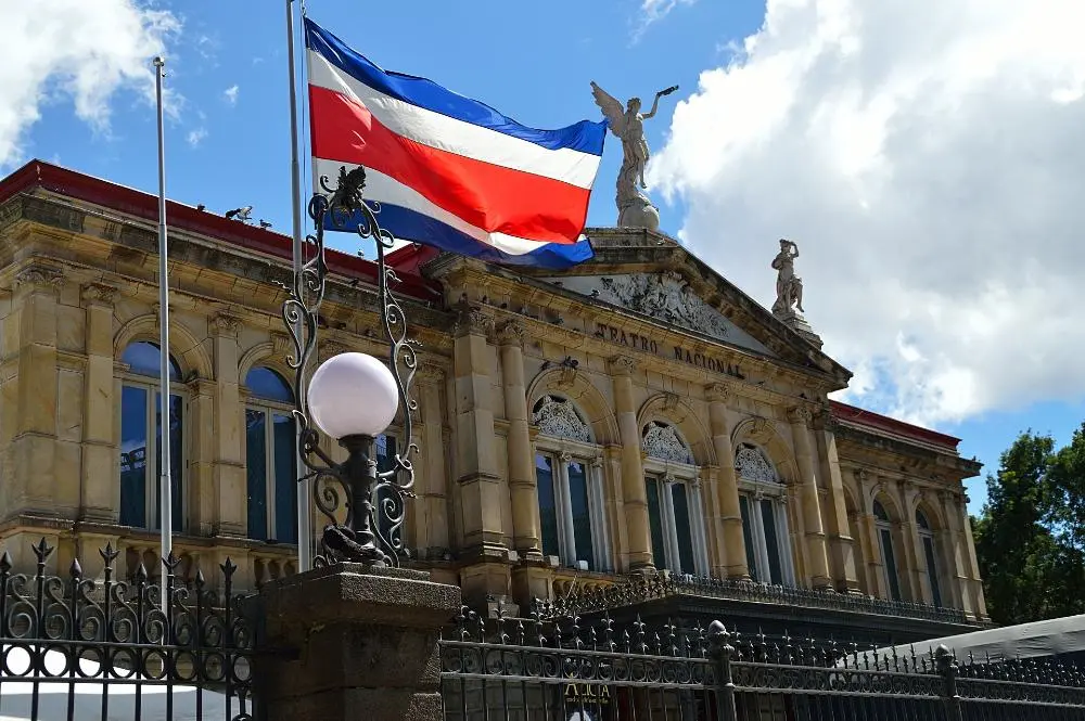 The image shows the National Theater of Costa Rica, a grand building with ornate architecture, including statues and decorative elements. The Costa Rican flag waves prominently in the foreground. The sky is partly cloudy.