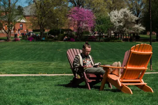 A person is seated on a wooden chair on a grassy lawn, reading. They are surrounded by blooming trees and more empty chairs. The background features a brick building and people walking under a sunny sky.