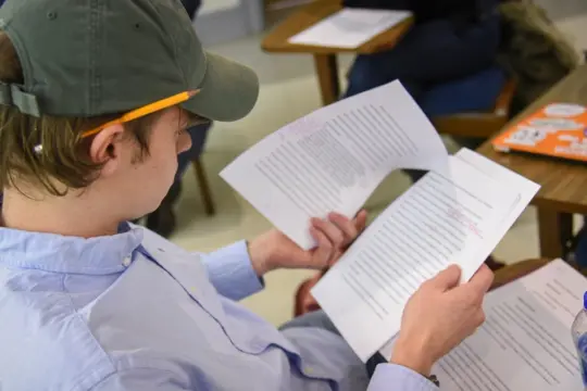 A person in a cap reviews paper documents with handwritten notes in a classroom setting. A pencil is tucked behind their ear, and there is a laptop with stickers on a desk nearby.