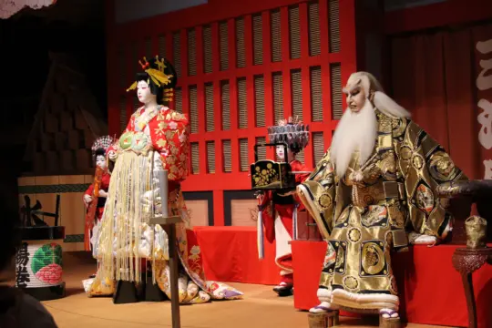 A traditional Japanese kabuki performance, perfect for enthusiasts of Asian Studies, features actors in vibrant costumes and makeup on stage. The scene includes a central figure in an elaborate red and gold outfit, surrounded by others in equally detailed attire.