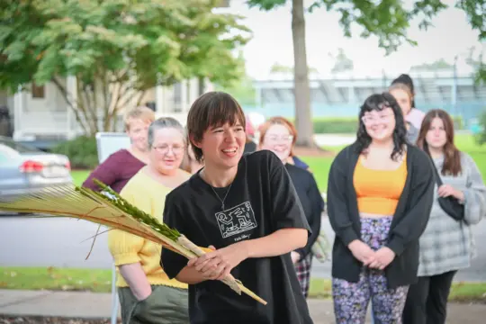 A young person smiling and holding palm leaves stands in front of a small group of people outdoors during a Jewish & Israel Studies event. The group looks on and smiles, with trees and a street visible in the background.