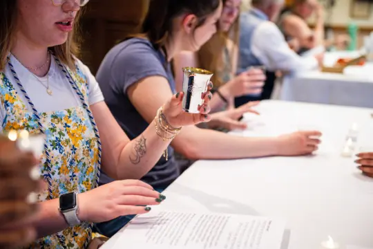Several people sit at a table holding small, silver goblets. One woman in a floral dress looks at her goblet, with papers and candles—suggesting a Jewish & Israel Studies gathering—on the table in front of the group.