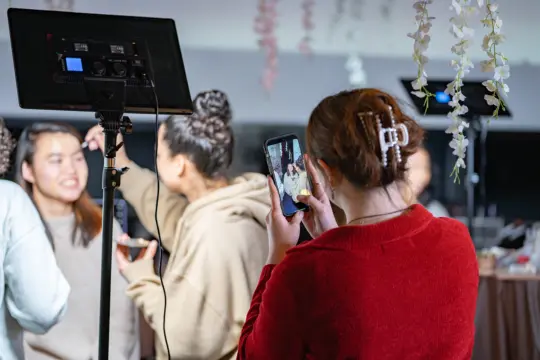 Person in red sweater takes a photo with a smartphone at an indoor setting with floral decorations. A group of people are gathered, some appear to be taking selfies. Studio lighting equipment is visible in the foreground.