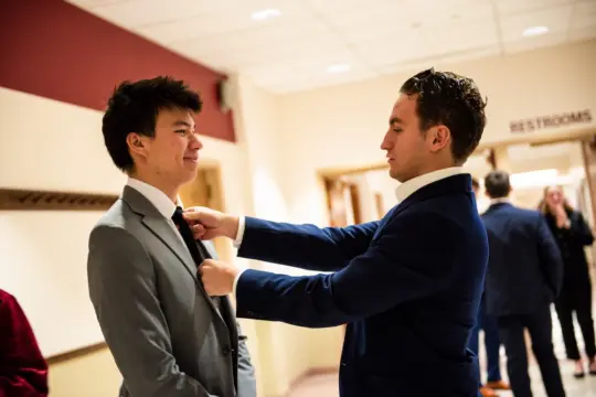 Two men in suits in a brightly lit hallway. One adjusts the tie of the other, who smiles. A 